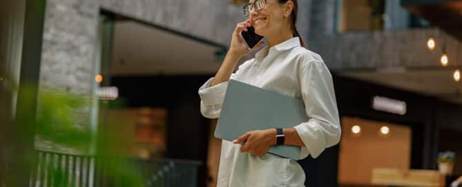 Franchise Recruitments woman talking phone with client while walking in office hall with laptop.