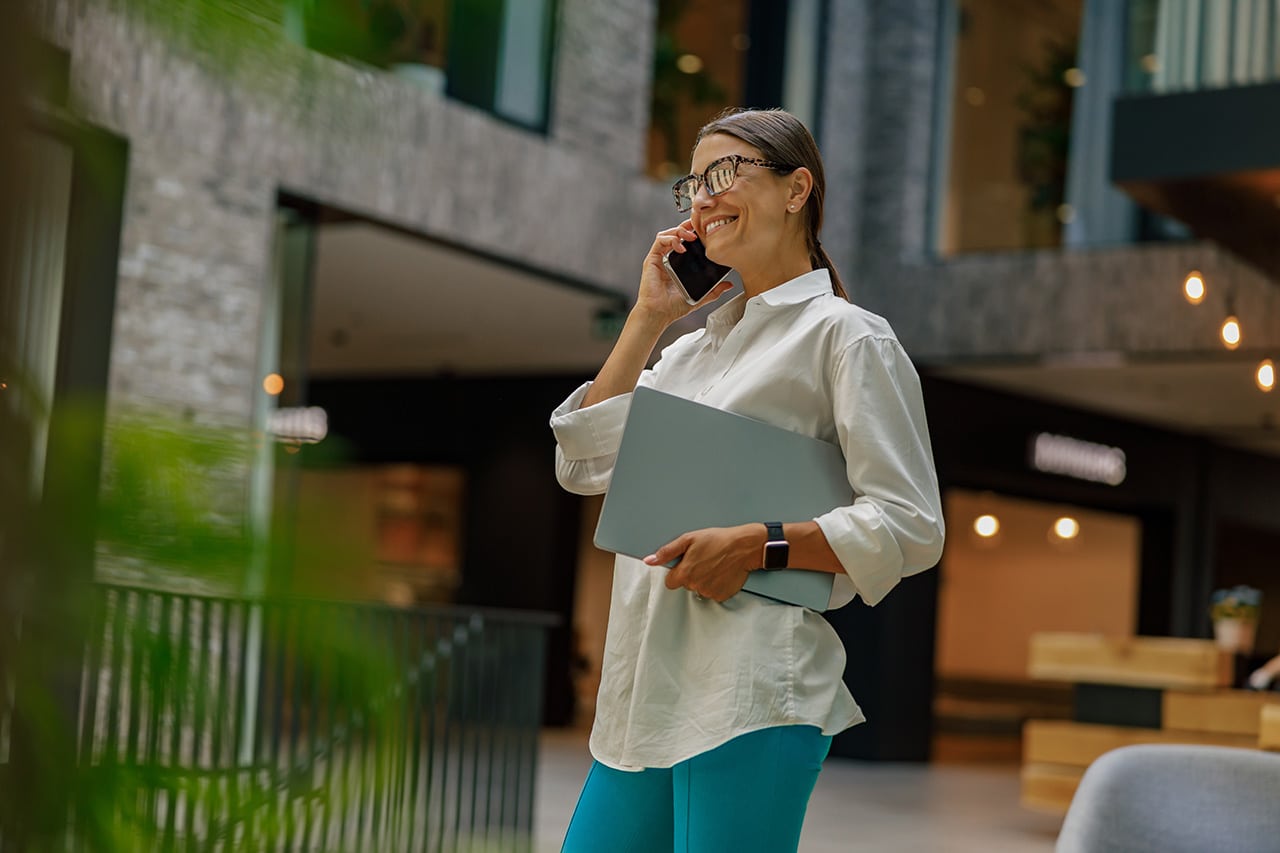 Franchise Recruitments woman talking phone with client while walking in office hall with laptop.